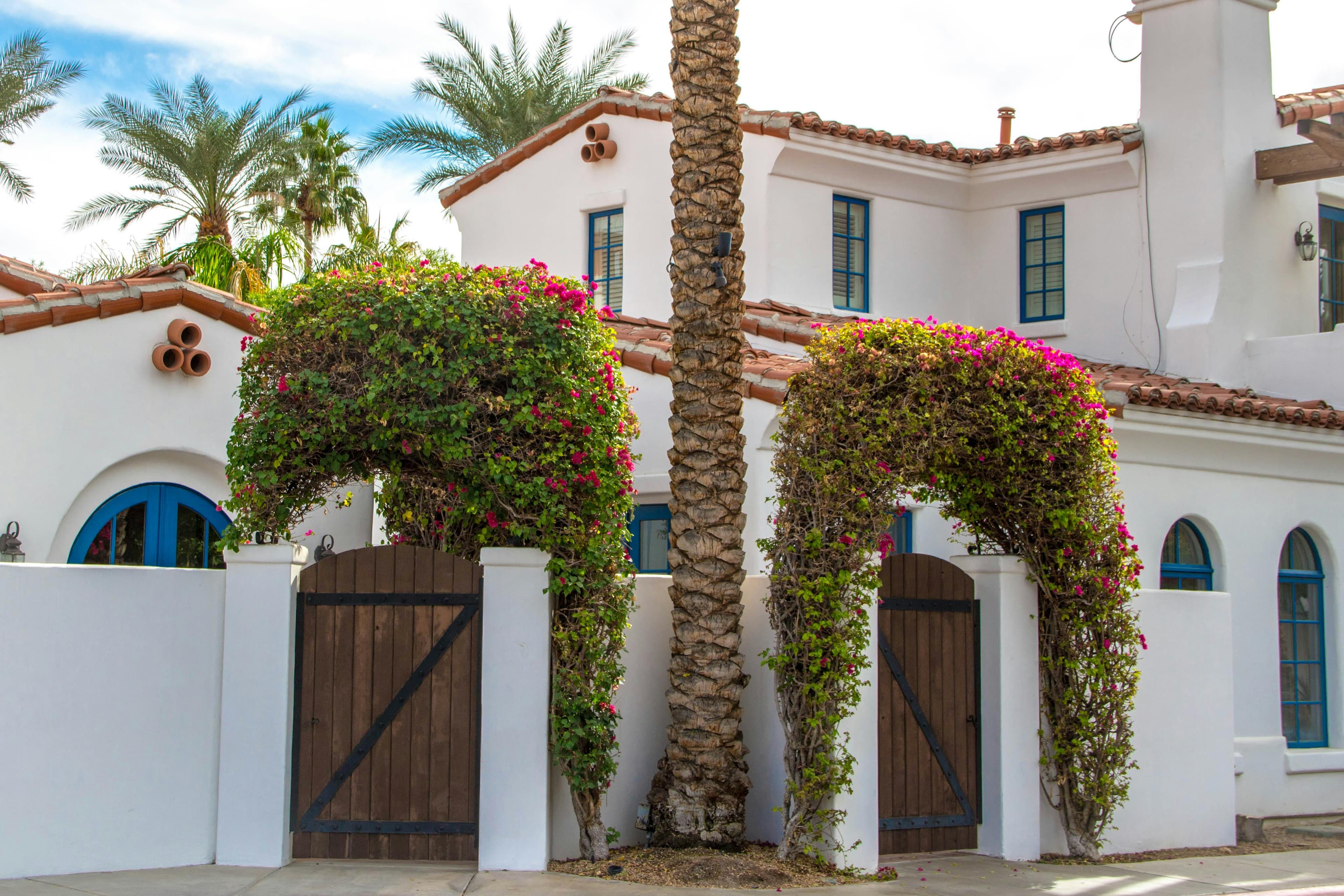 Mediterranean-style house with white walls, blue windows, and red-tiled roof. Bougainvillea arches over wooden gates, palm trees in view.
